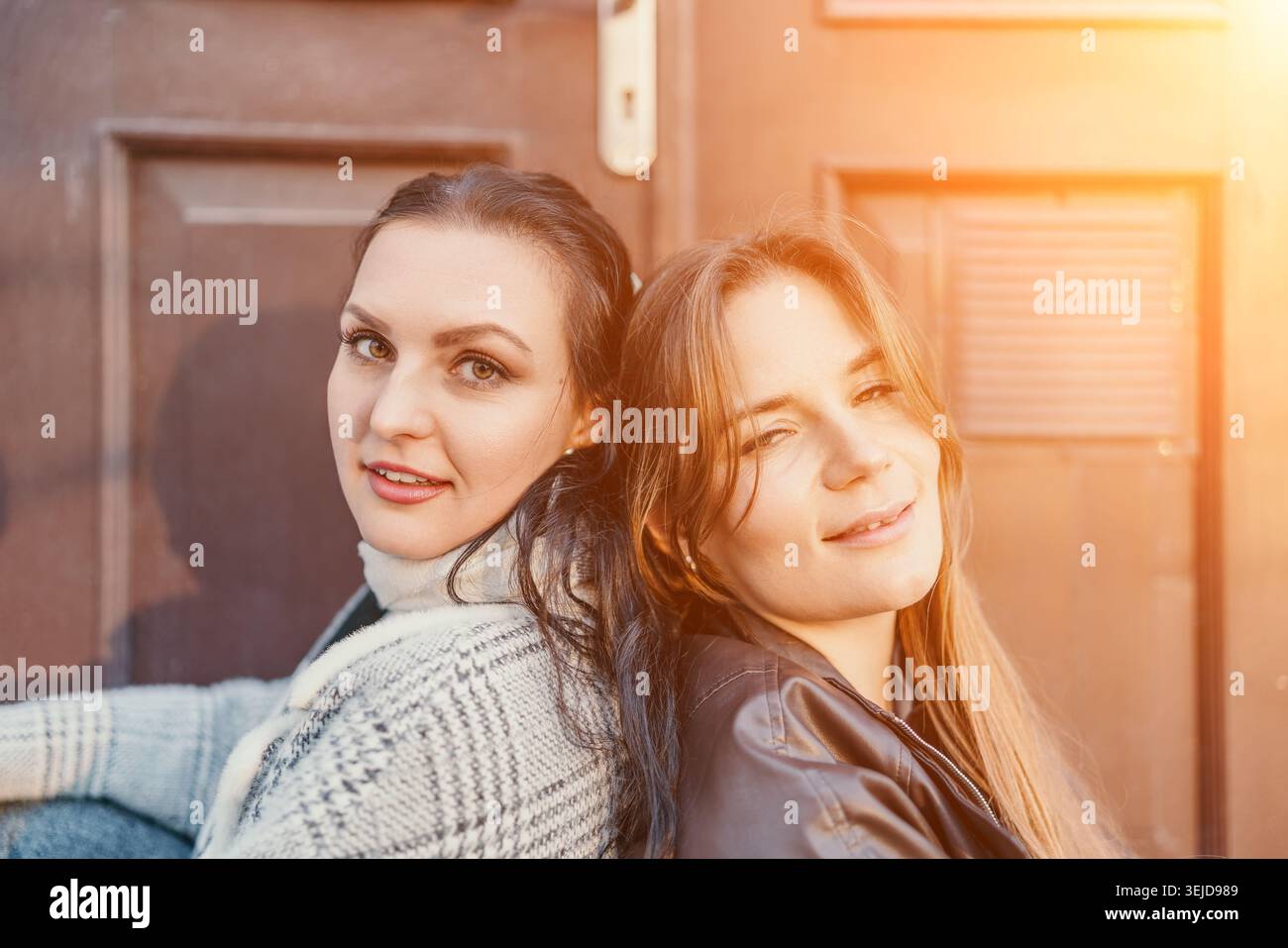 Stile di vita degli amici al tramonto due giovani donne sorridenti si appoggiano schiena a schiena contro una porta di legno illuminata dalla calda luce del sole dorata Foto Stock