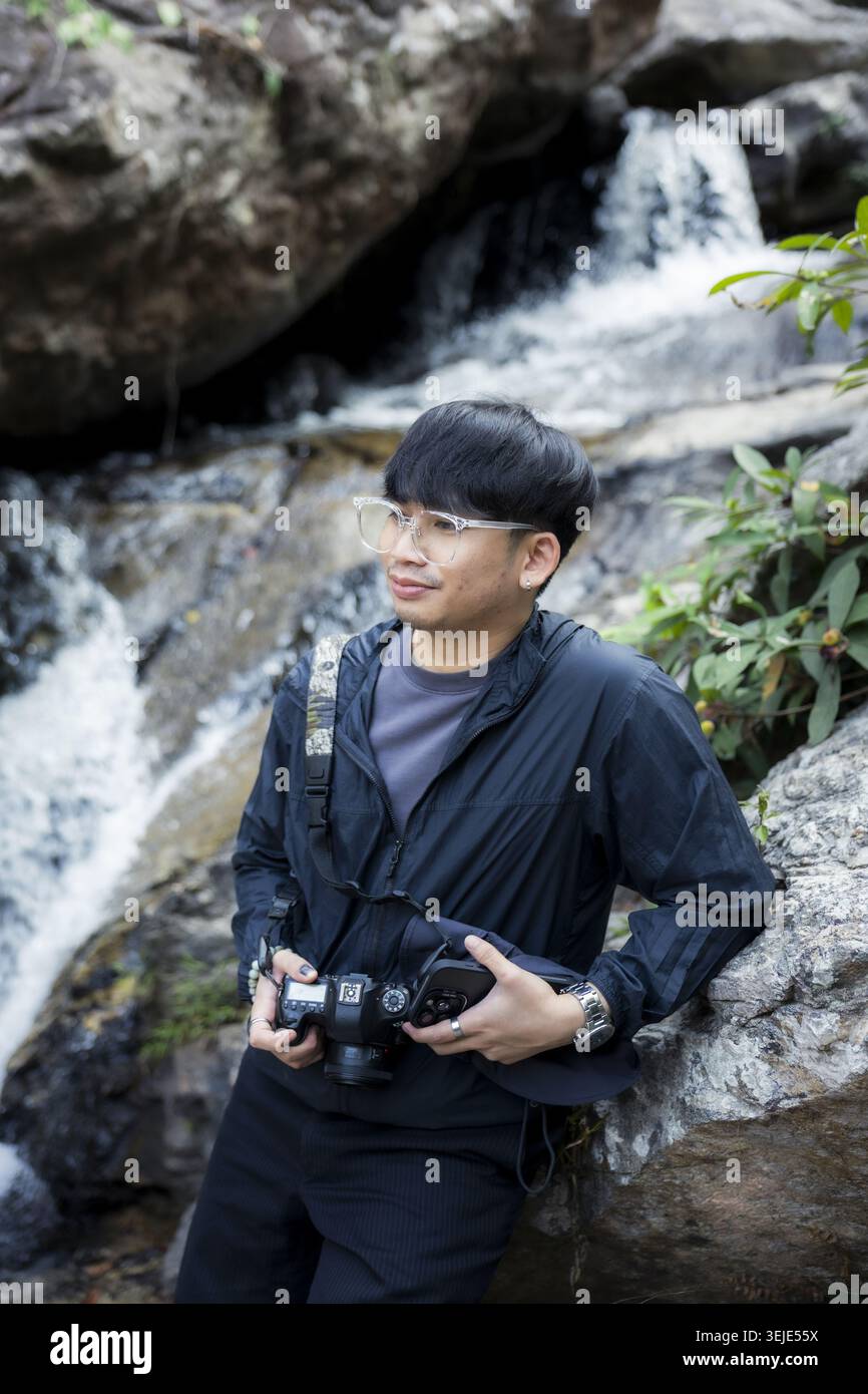 Giovane fotografo asiatico dall'espressione pacifica e felice tenendo la macchina fotografica in piedi accanto alla cascata rocciosa naturale nella foresta verde godendo della natura all'aperto Foto Stock