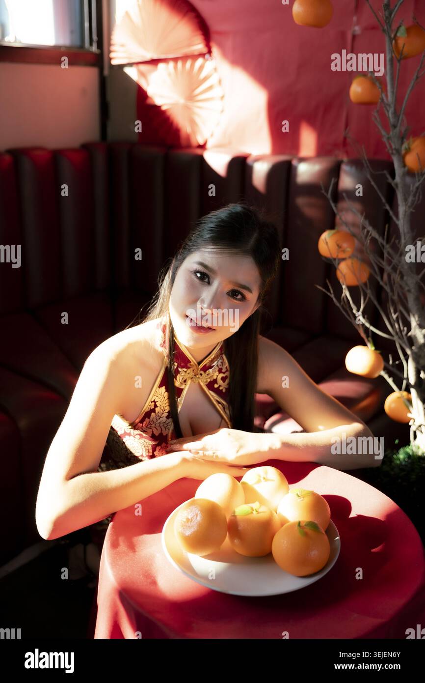 Bella ed elegante donna asiatica in cheongsam rosso ha un'espressione serena durante la celebrazione del capodanno cinese. ritratto con arance che simboleggiano la fortuna e. Foto Stock