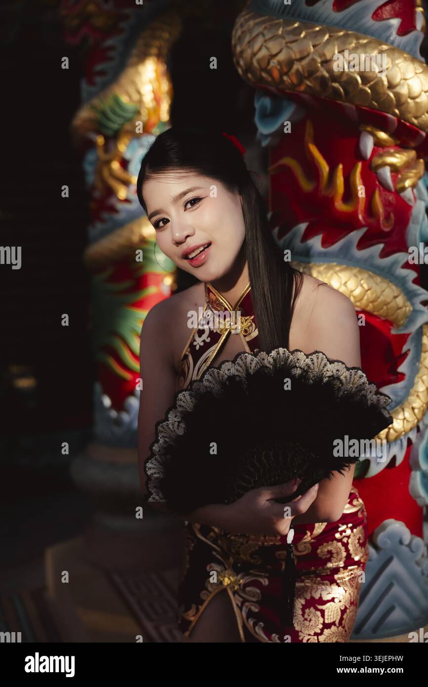 Bella donna asiatica con tradizionale abito cinese qipao rosso e oro, ha un fan nero. Si trova graziosamente vicino alla statua del drago dorato nel tempio, con Foto Stock