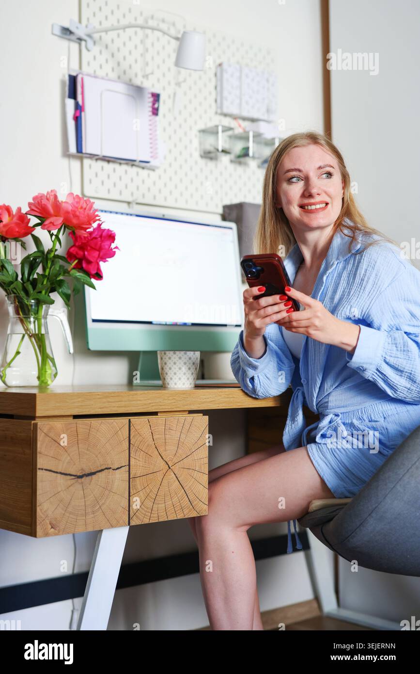 Donna sorridente seduta alla scrivania con smartphone e peonie, moderno ritratto dell'ufficio domestico per la comunicazione e i temi dello stile di vita digitale. Foto Stock
