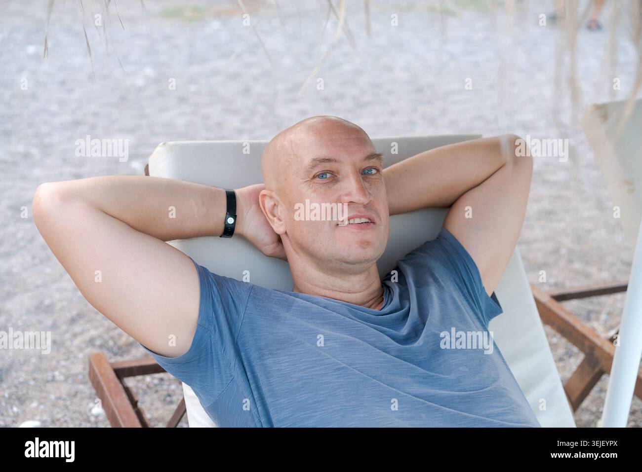 L'uomo caucasico calvo con occhi blu sorride leggermente rilassandosi con le braccia dietro la testa sul lettino in spiaggia. Adatto per soggiorni benessere e spiaggia Foto Stock