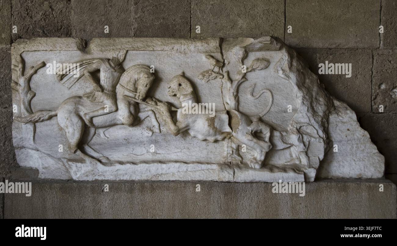 Frammento di una targa di marmo raffigurante una scena di caccia. Un cavaliere, rivestito con un mantello e un pettorale su una tunica corta, armato di lancia, affronta una leonessa che si affonda verso di lui. Sul margine destro del rilievo, appena visibile, un cane da caccia attacca la leonessa da dietro, mentre una seconda figura umana con il braccio destro sollevato, probabilmente con una mazza, sta per colpire la bestia. Periodo tardo ellenistico. Trovato negli scavi del Tetrapylon romano nella città di Rodi. Museo archeologico di Rodi, Grecia. Foto Stock