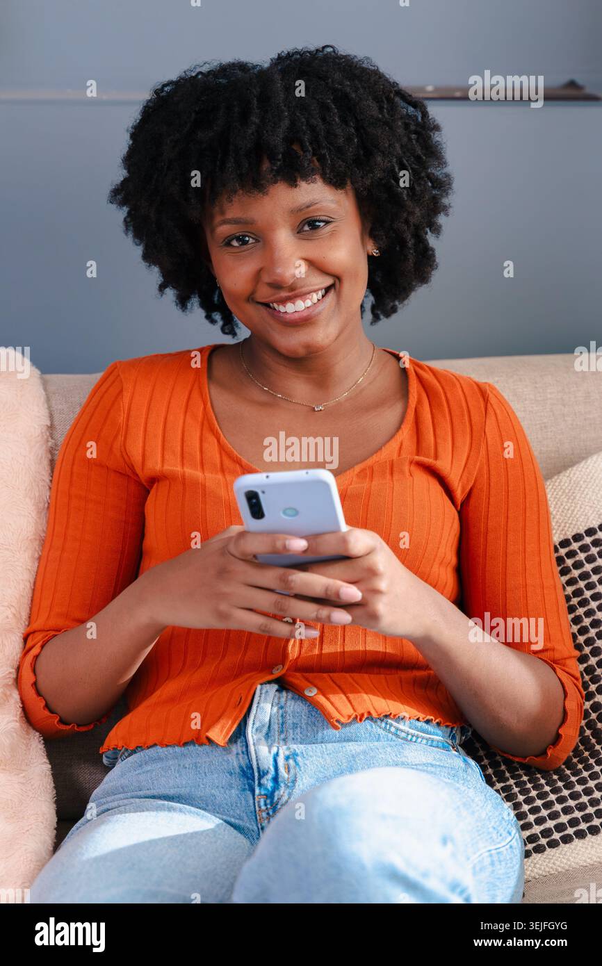 Ritratto verticale allegra donna nera che ride mentre usa uno smartphone sul divano di casa Foto Stock
