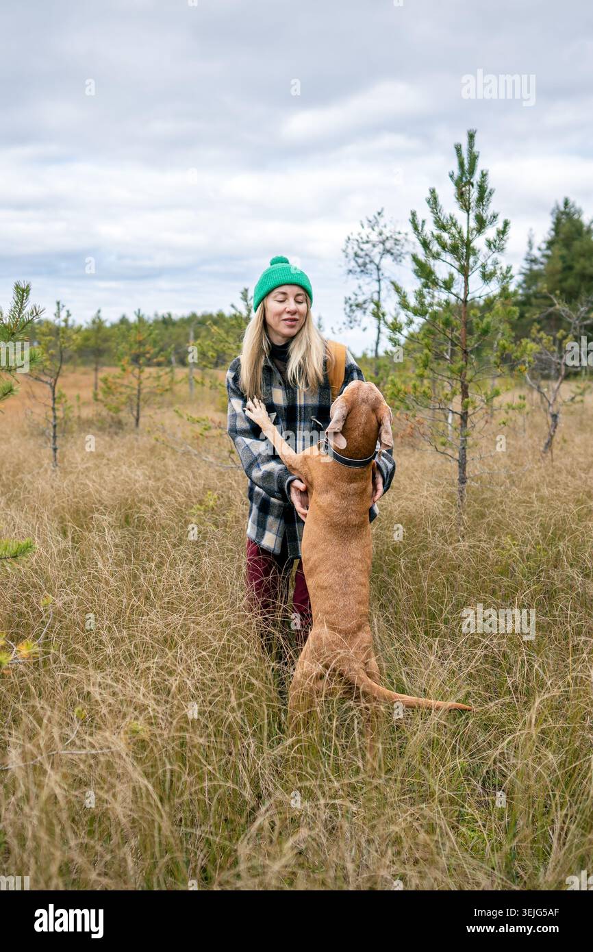 Donna proprietaria di un animale domestico che gioca con il cane all'aperto, in piedi sul prato autunnale di erba seghettata nella zona umida autunnale. Foto Stock
