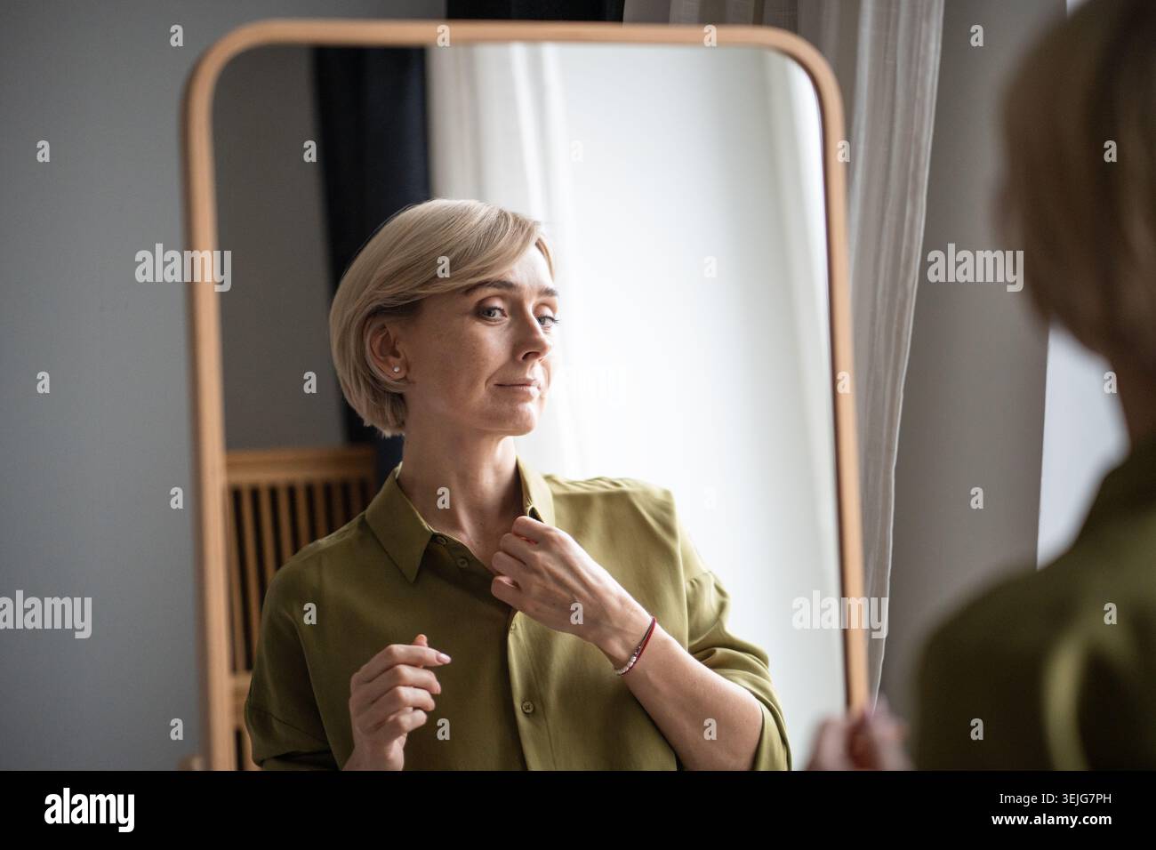 Una donna bionda di mezza età che si prepara per l'appuntamento, controlla la prospettiva davanti allo specchio a casa. Foto Stock