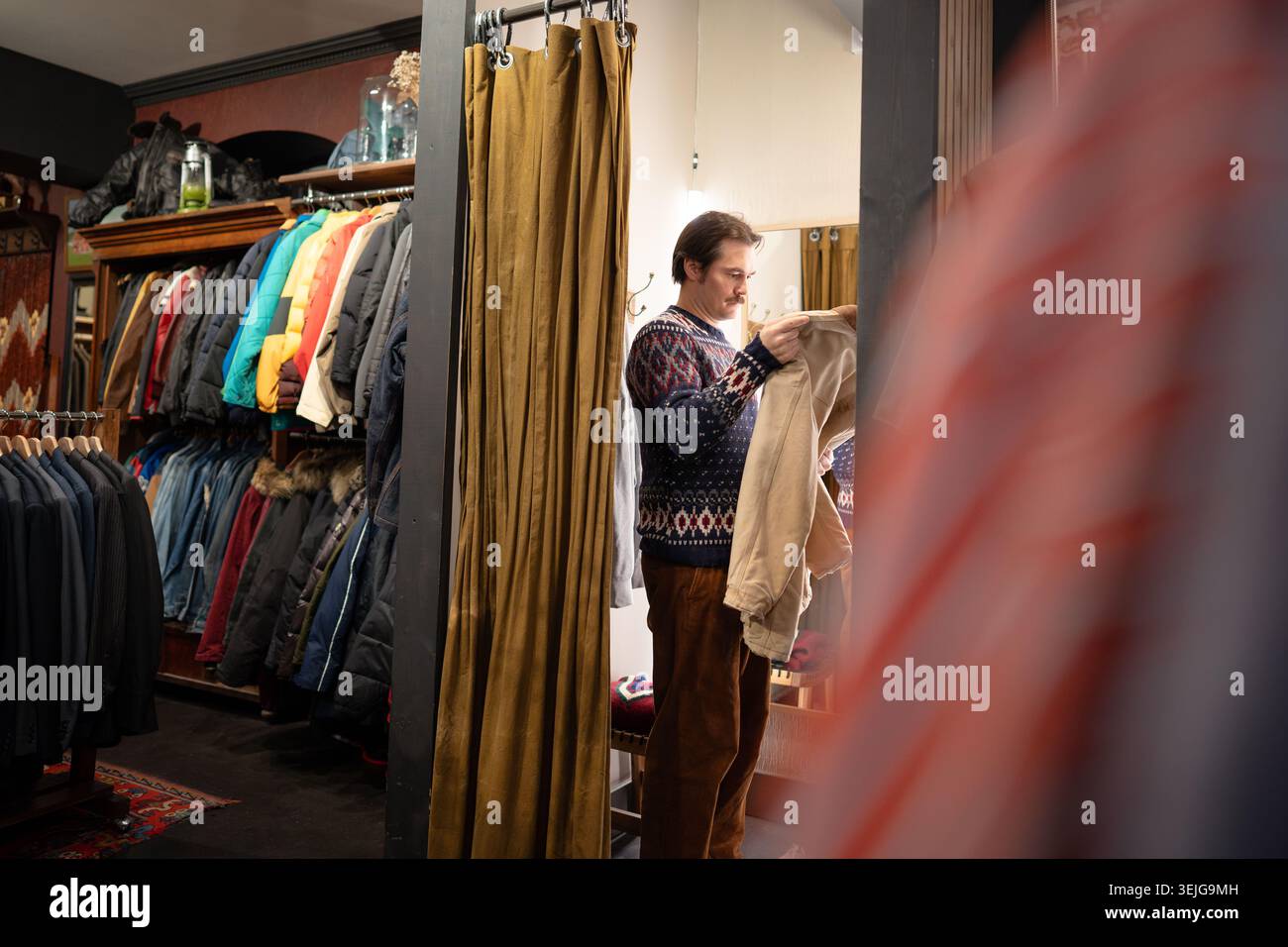 Un uomo elegante che acquista abiti vintage in un negozio di seconda mano, provando una giacca in camerino. Foto Stock