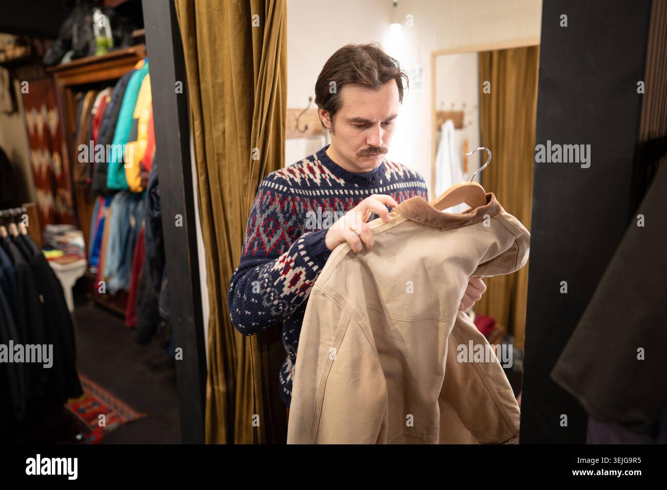 Uomo affascinante che sceglie abiti di seconda mano in boutique vintage, controllando la qualità degli outerwear. Foto Stock
