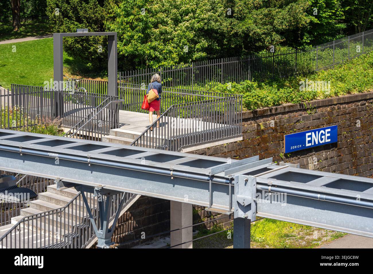 Zurigo, Svizzera - 21 maggio 2026: Donna che cammina su un sentiero accanto a un ponte di acciaio, scale, recinzione e alberi verdi. Stazione ferroviaria urbana di Zurigo Enge. Foto Stock