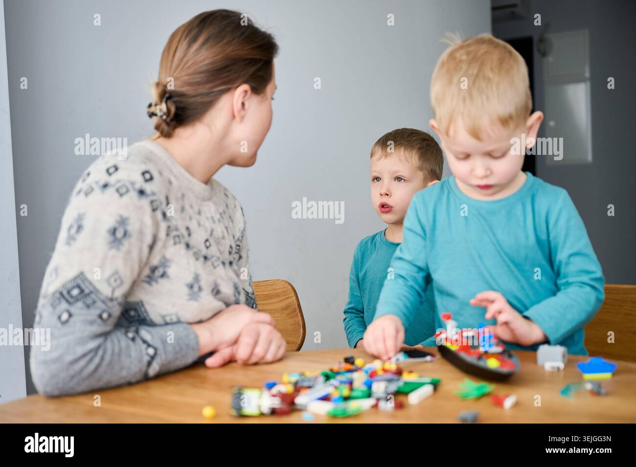 Ragazzo che gioca con i blocchi di costruzione mentre un altro ragazzo parla con la madre. Sedersi a tavola a casa, interagire con la famiglia, giocare e comunicare con i bambini Foto Stock