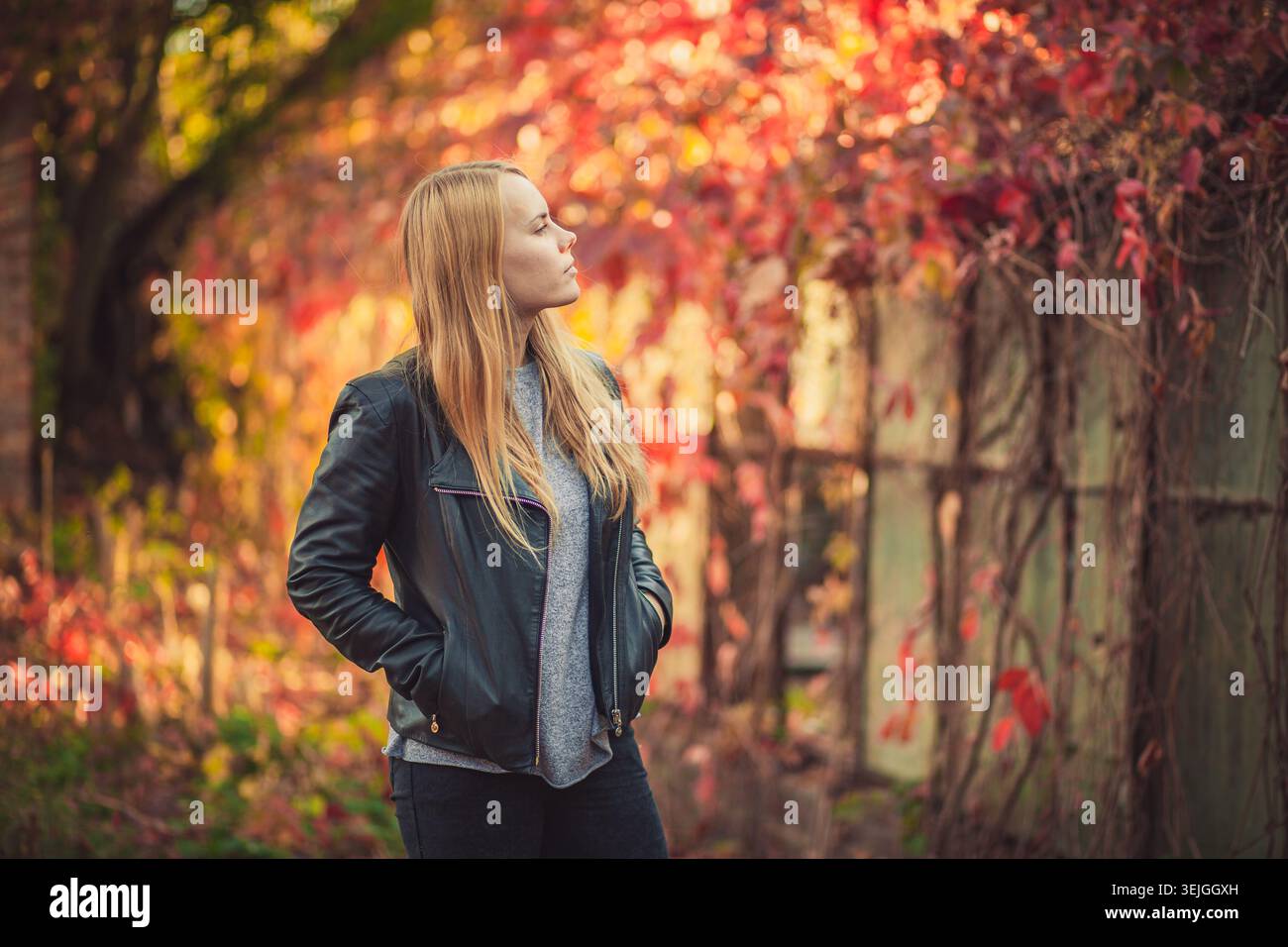 Giovane donna premurosa con capelli biondi sta di profilo. Indossa un abbigliamento casual autunnale, con le mani nelle tasche della sua giacca nera in pelle Foto Stock