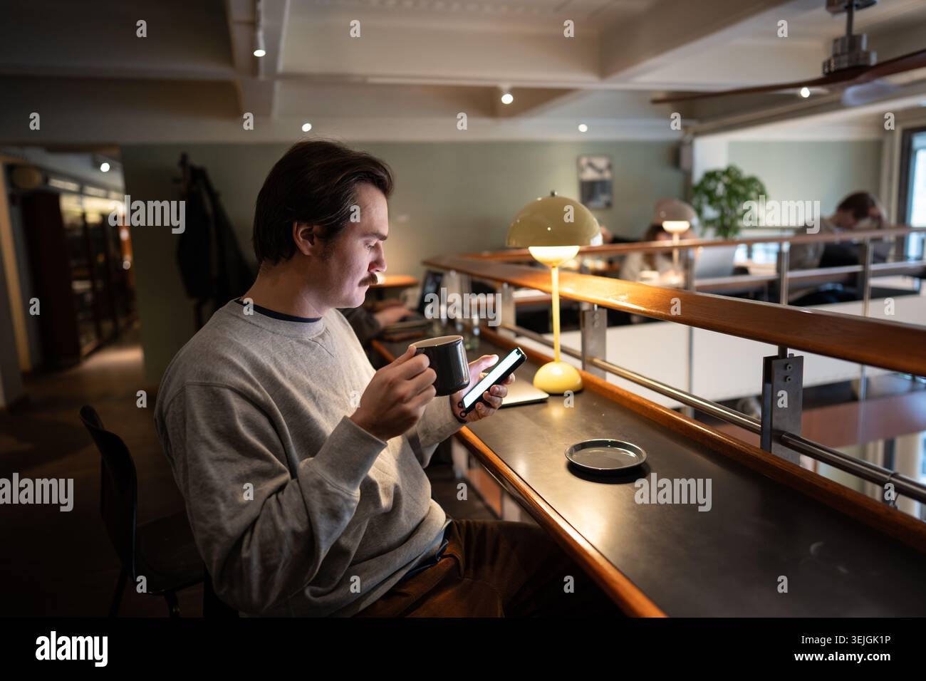 Uomo rilassato e premuroso che beve caffè e controlla i messaggi sullo smartphone nello spazio della caffetteria della libreria. Foto Stock