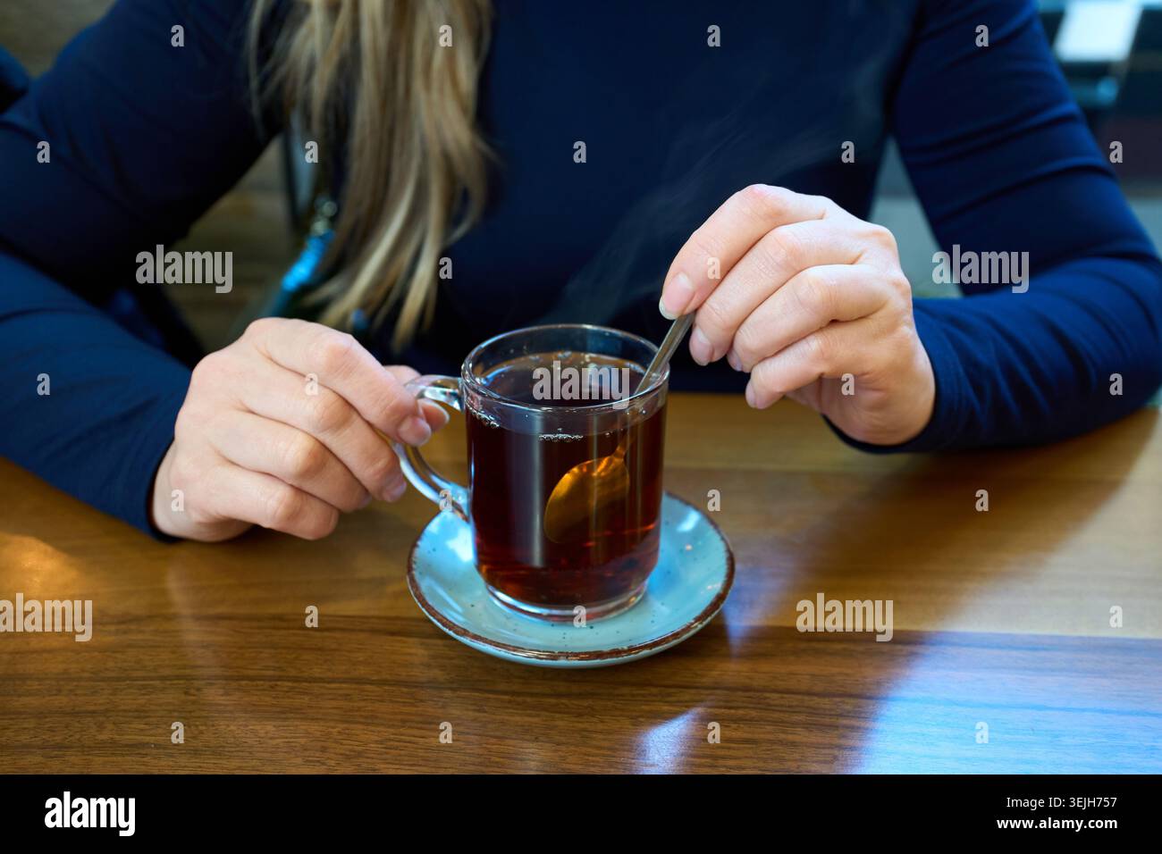 Una donna che mescola tè nero caldo con un cucchiaio in una tazza di vetro trasparente su un tavolo di legno all'interno di un caffè. Foto Stock