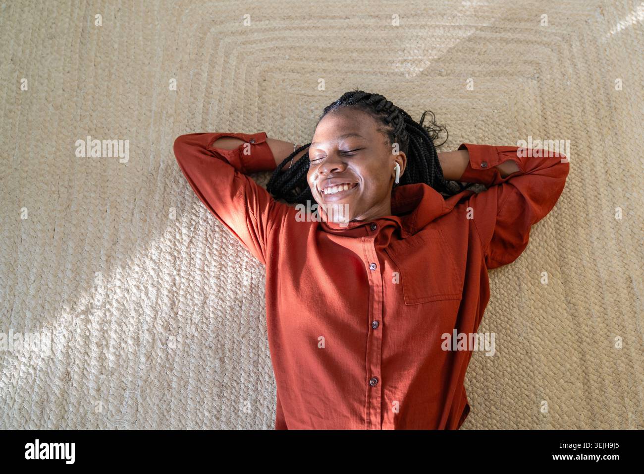Una donna afroamericana spensierata e felice, sdraiata sul pavimento, che si rilassa ascoltando la musica preferita in baccelli. Foto Stock