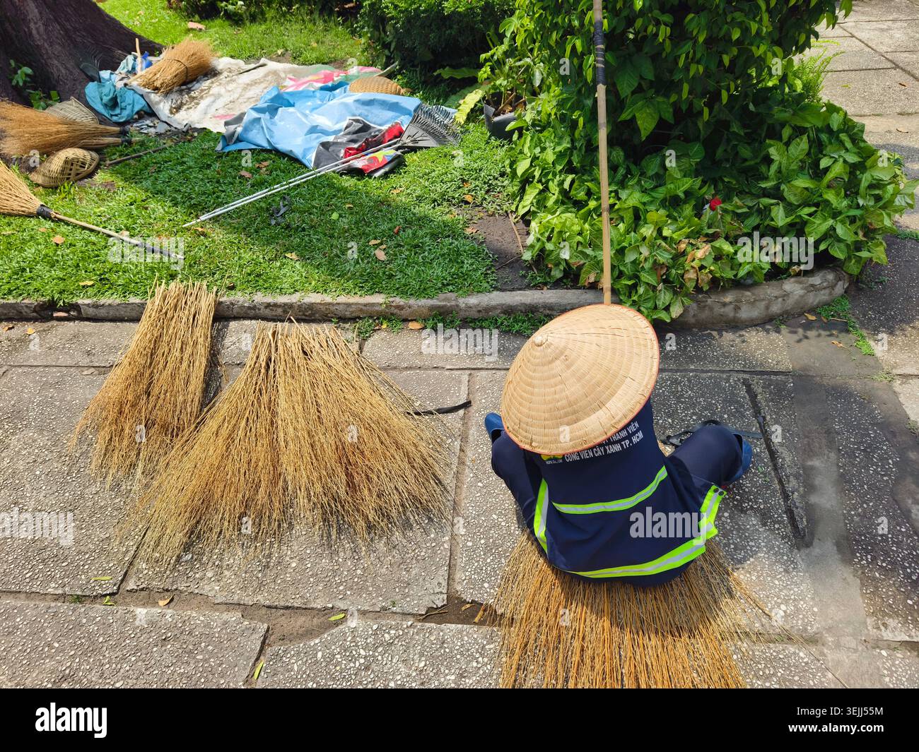 Una donna che indossa un cappello conico e un'uniforme riflettente che lavora all'aperto con le tradizionali scope fatte a mano realizzate in erba secca. HCMC, Vietnam Foto Stock