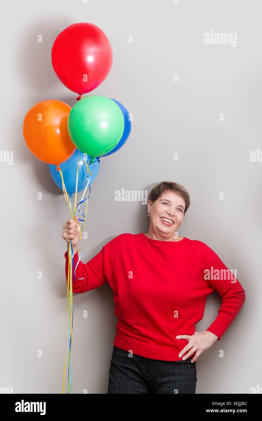 Signora anziana felice che tiene regali e palloncini multicolori che guardano la macchina fotografica e sorridono in posa intorno a un muro. Natale. B Foto Stock
