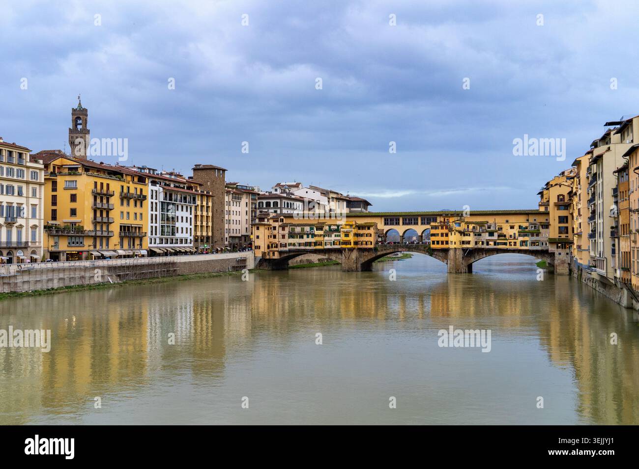 L'iconico Ponte Vecchio di Firenze, in Italia, fiancheggiato da negozi e che si riflette sull'Arno sotto un cielo nuvoloso Foto Stock