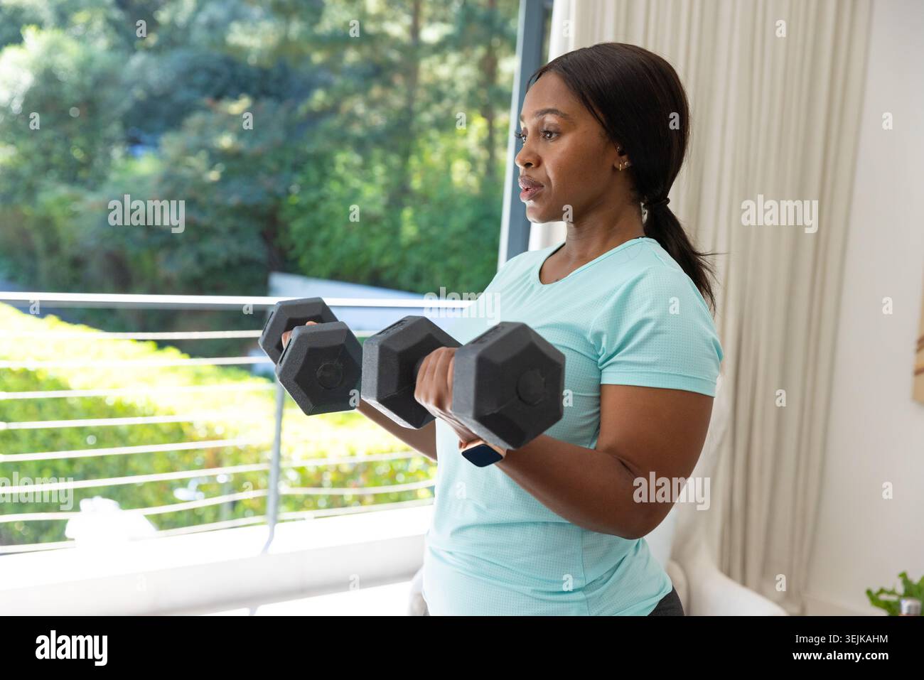 Donna africana che solleva manubri esagonali neri con una leggera T-shirt da atletica Aqua vicino a una porta in vetro scorrevole Foto Stock