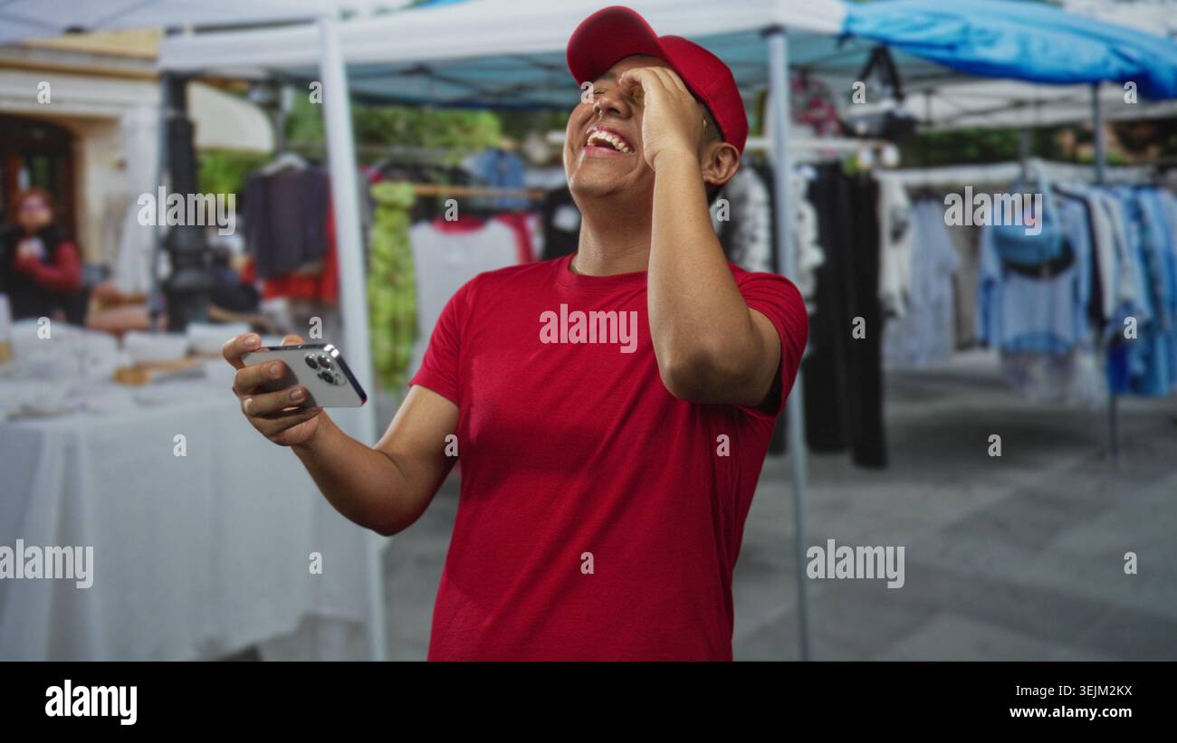 Uomo che tiene lo smartphone con entrambe le mani nelle bancarelle del mercato di strada, indossa un berretto rosso e una maglietta rossa, ridendo e sorridendo; divertimento di gioco. Foto Stock