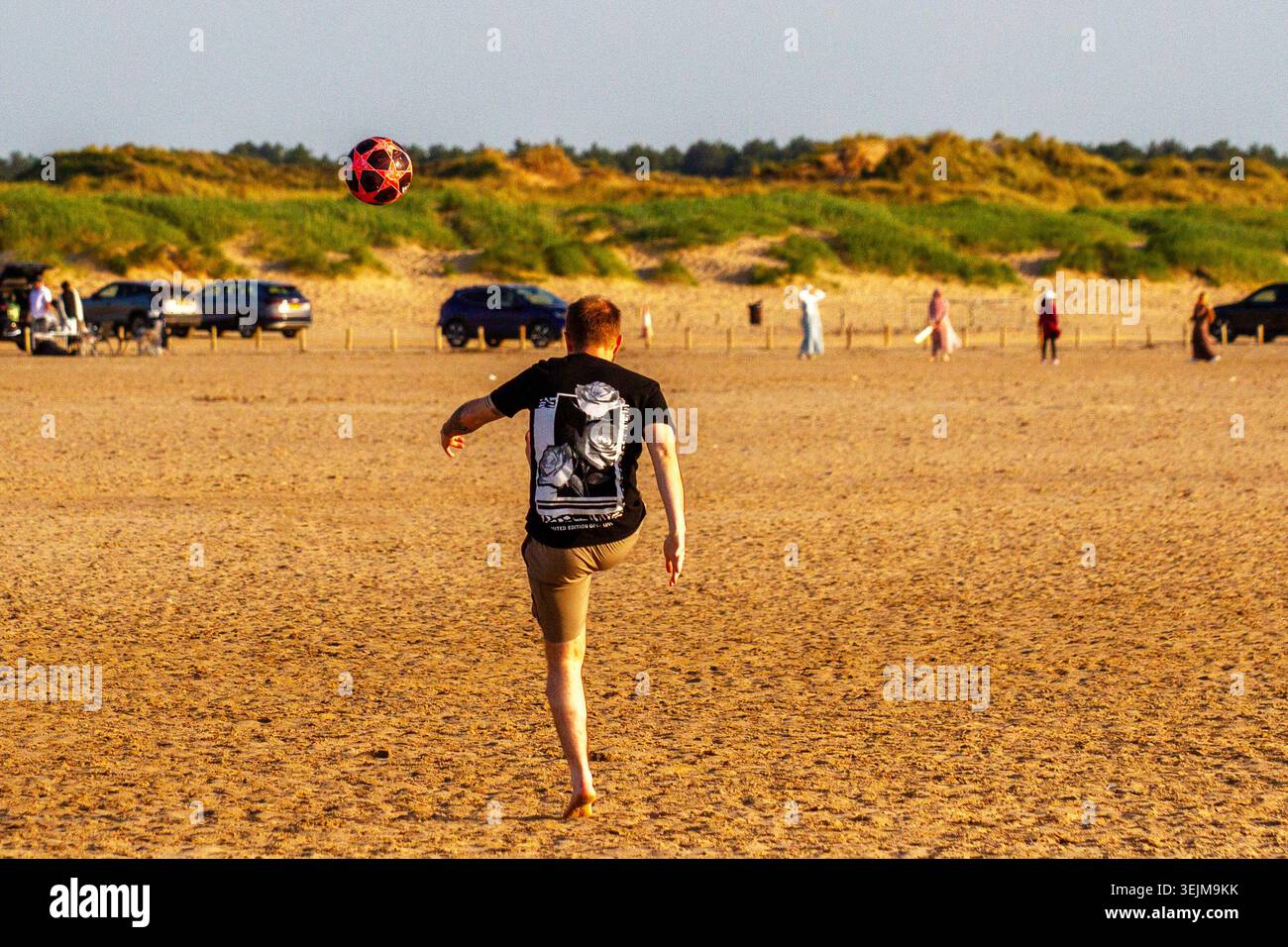 Vista posteriore di un giovane adulto che gioca con un pallone a fantasia su un'ampia spiaggia sabbiosa con dune erbose e auto parcheggiate sullo sfondo. Foto Stock