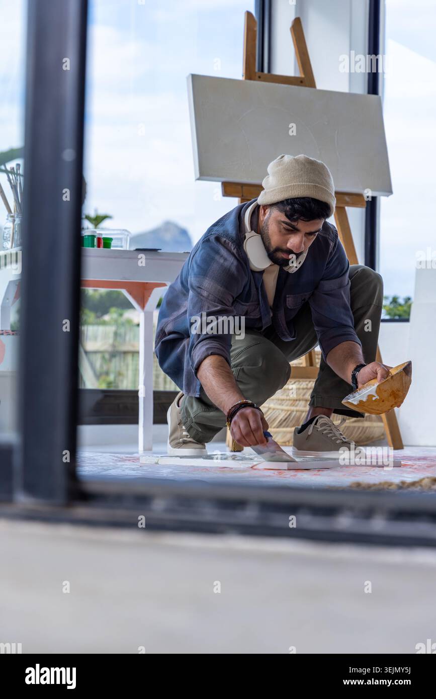 Artista maschile accovacciato su tela da pavimento con tavolozza, indossando berretto e cuffie. Cavalletto, pennello, pittura, atelier, Foto Stock