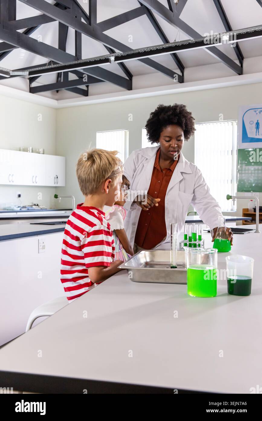 Istruttore afroamericano che indossa un camice da laboratorio bianco che insegna a due bambini nel laboratorio scolastico con i becher. Classe, scienza, glasswa Foto Stock