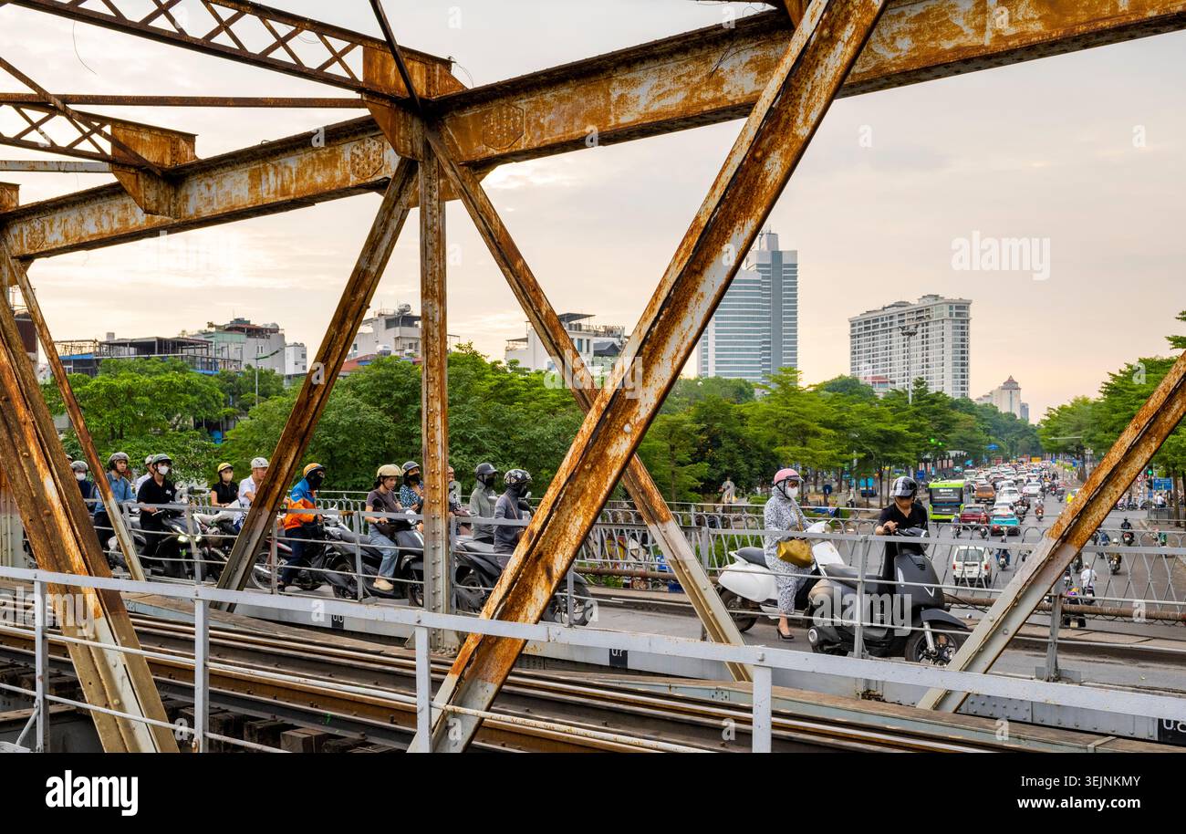 Vietnam-11 giugno 2025: I pendolari nei giorni feriali in moto viaggiano su stretti sentieri, su entrambi i lati del vecchio ponte coloniale francese in acciaio, che attraversa la R di Hanoi Foto Stock
