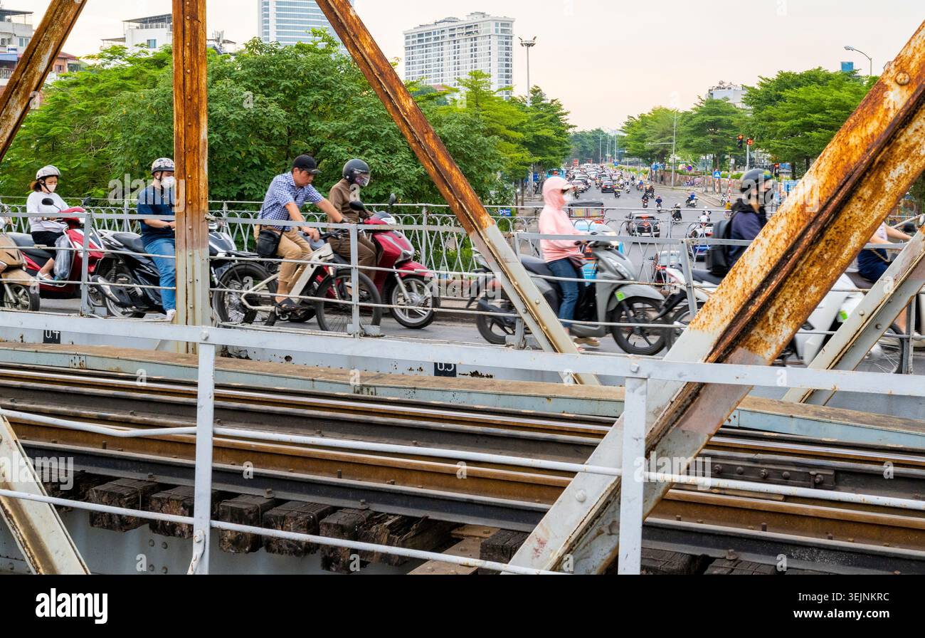Vietnam-11 giugno 2025: I pendolari nei giorni feriali in moto viaggiano su stretti sentieri, su entrambi i lati del vecchio ponte coloniale francese in acciaio, che attraversa la R di Hanoi Foto Stock