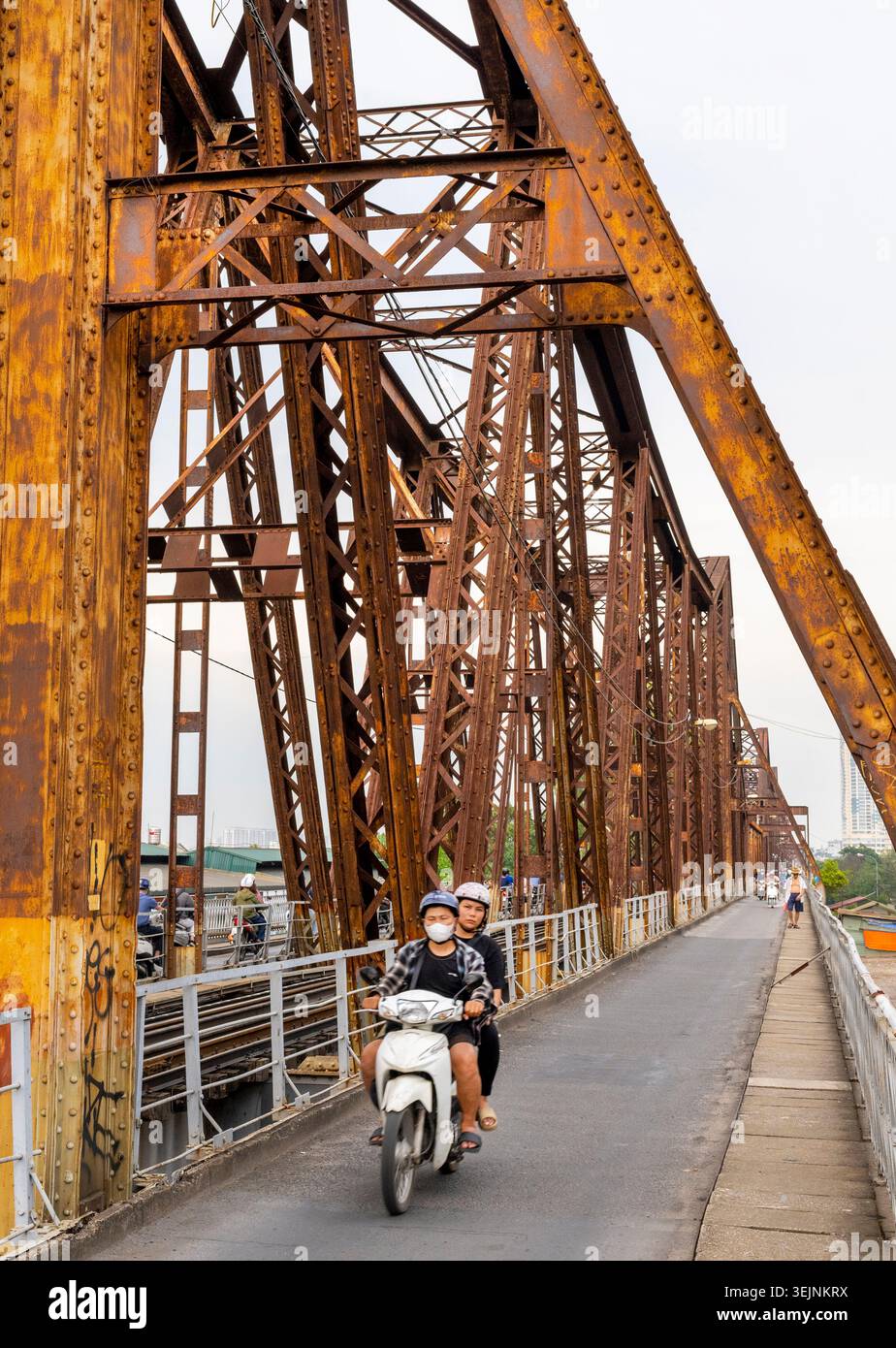 Vietnam-11 giugno 2025: I pendolari nei giorni feriali in moto viaggiano su stretti sentieri, su entrambi i lati del vecchio ponte coloniale francese in acciaio, che attraversa la R di Hanoi Foto Stock