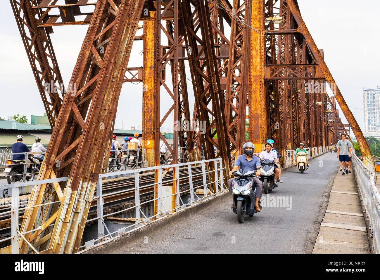 Vietnam-11 giugno 2025: I pendolari nei giorni feriali in moto viaggiano su stretti sentieri, su entrambi i lati del vecchio ponte coloniale francese in acciaio, che attraversa la R di Hanoi Foto Stock