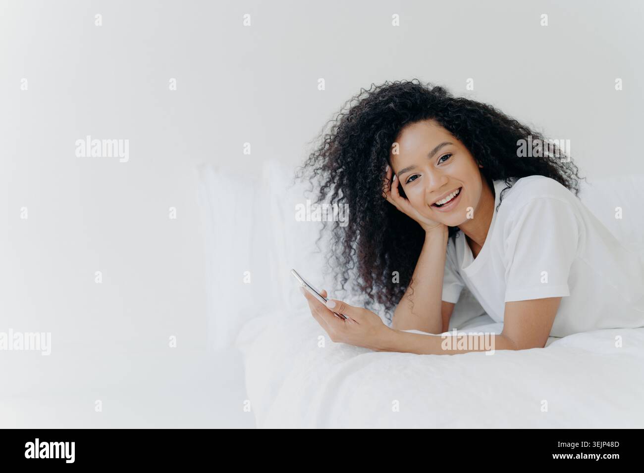 Felicissima, rilassata, riccia, giovane afroamericana gode del tempo libero a casa, si trova a letto con un moderno cellulare, usa il telefono Foto Stock