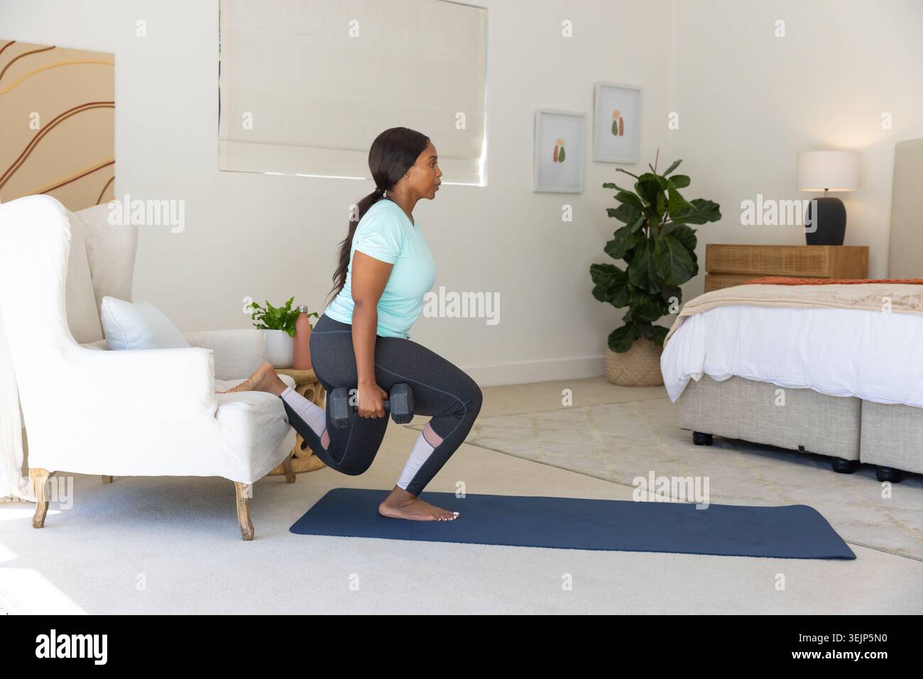 Adulto afroamericano che fa squat in camera da letto su un tappetino yoga che tiene i pesi su una sedia nei leggings Foto Stock