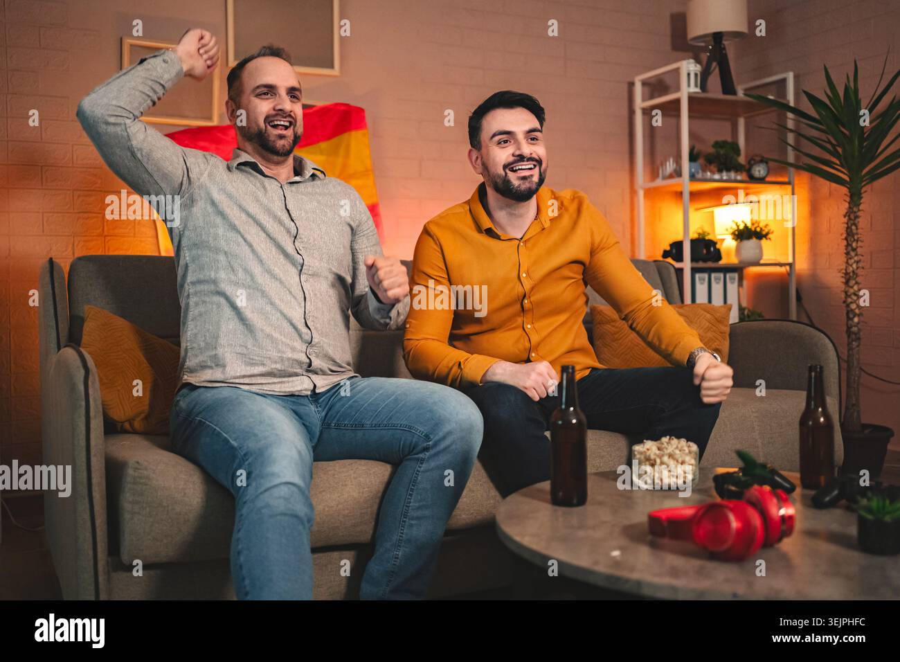 Gli amici celebrano una vittoria emozionante mentre guardano un evento sportivo a casa in una serata accogliente Foto Stock