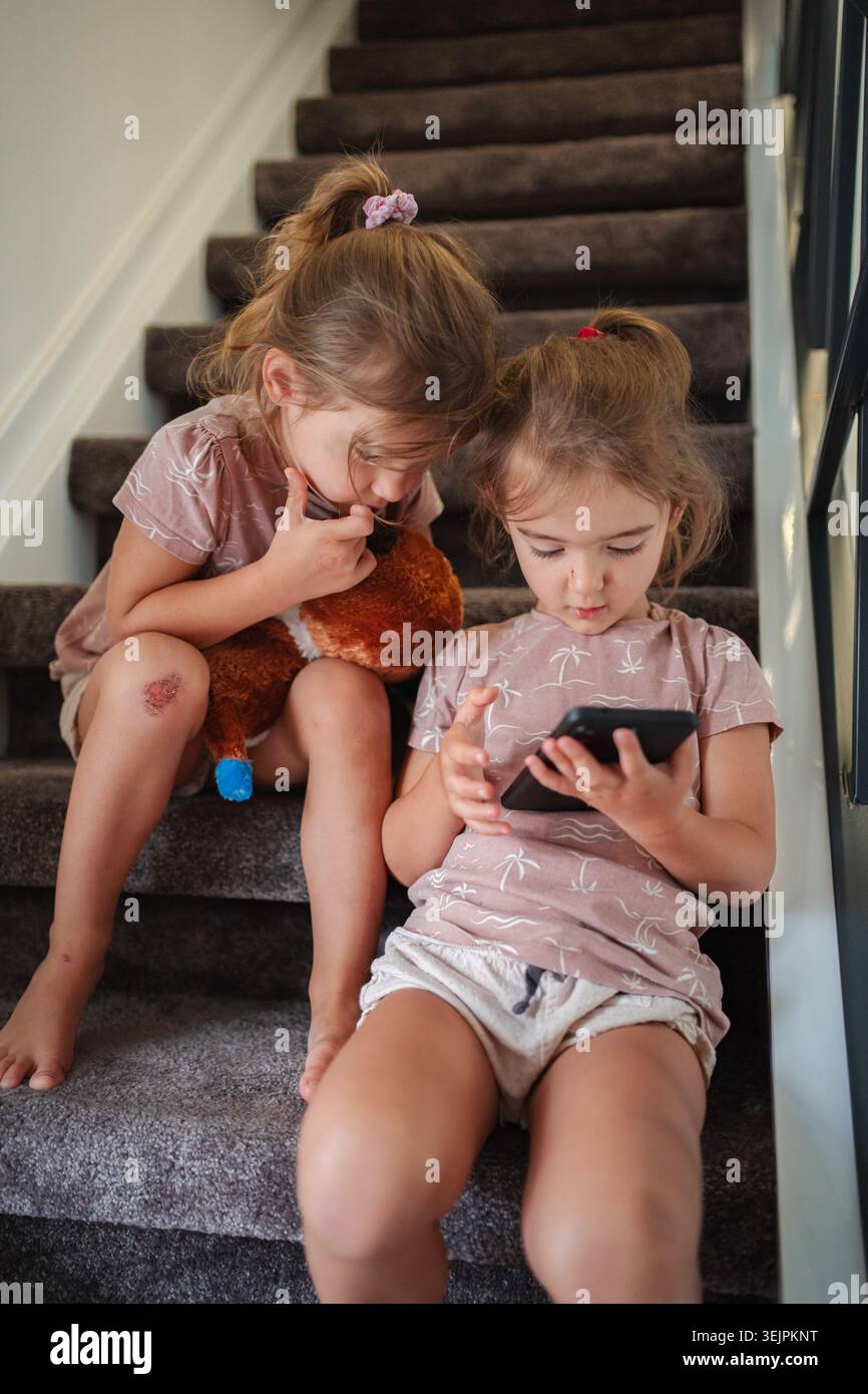 Due bambine in età prescolare che giocano con lo smartphone e l'orsacchiotto sulle scale Foto Stock