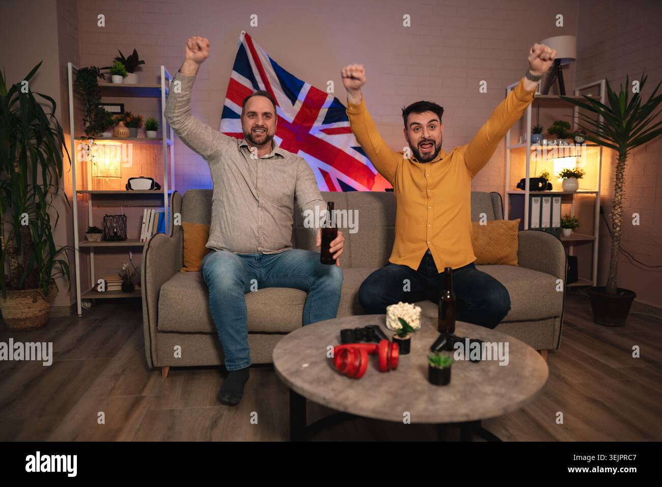 L'emozione riempie il salotto mentre gli amici celebrano una vittoria emozionante davanti a una bandiera del Regno Unito durante una vivace serata di gioco Foto Stock