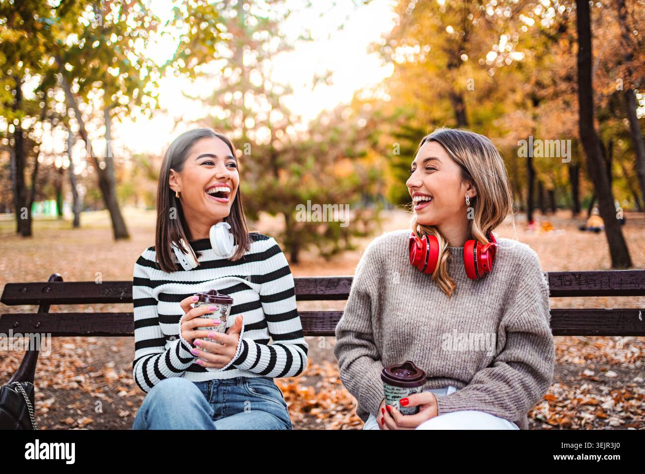 Le ragazze sorridono in autunno mentre sorseggiano bevande calde in un parco soleggiato Foto Stock