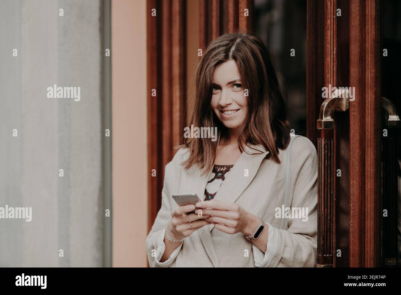Giovane donna sorridente che usa lo smartphone in piedi davanti all'ingresso dell'edificio Foto Stock