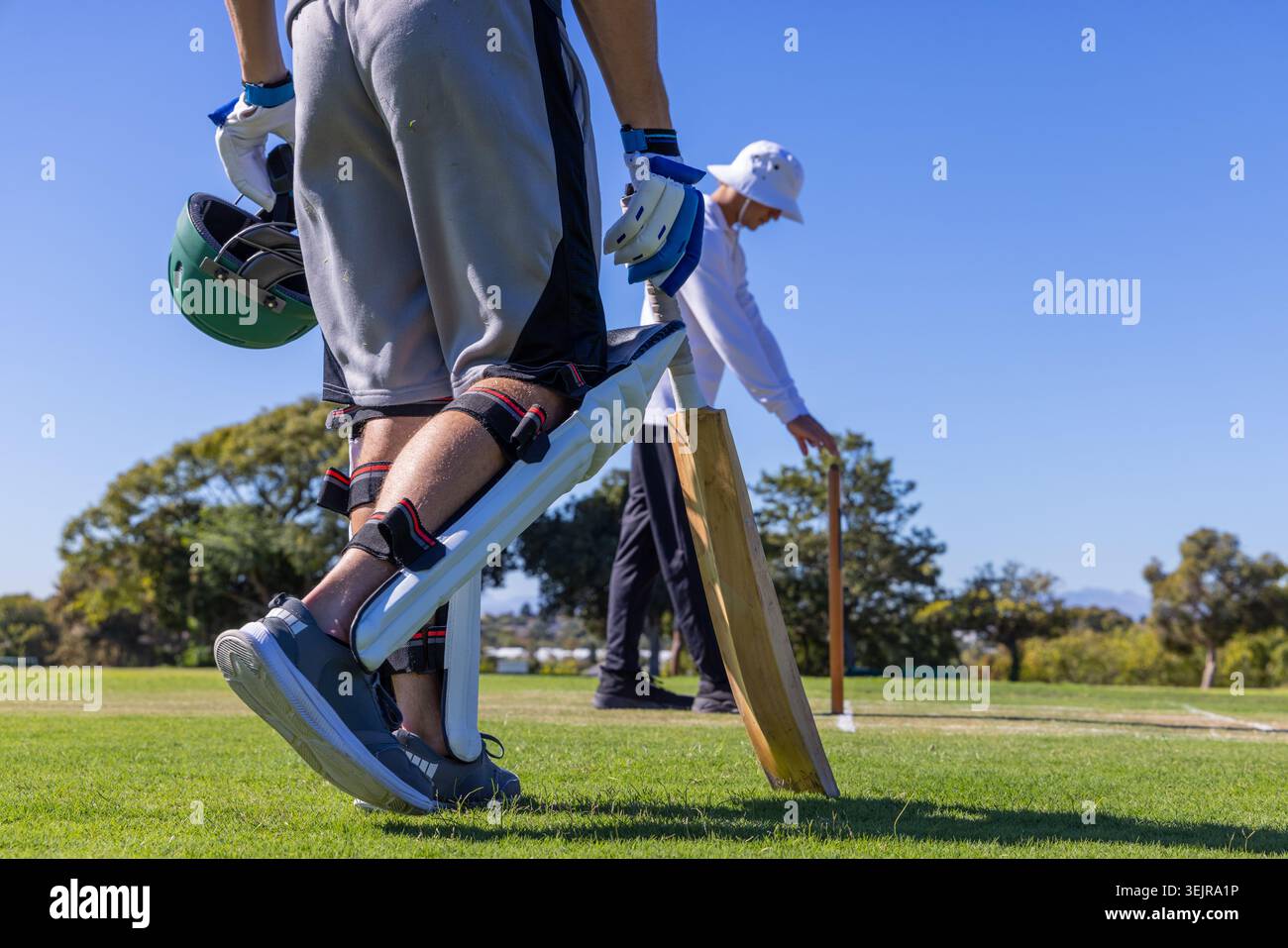 Due giocatori di cricket maschi in piedi sul campo di erba, batter in palle che tengono il casco e pipistrello vicino a ceppi. Cricket, all'aperto, soleggiato, diurno Foto Stock