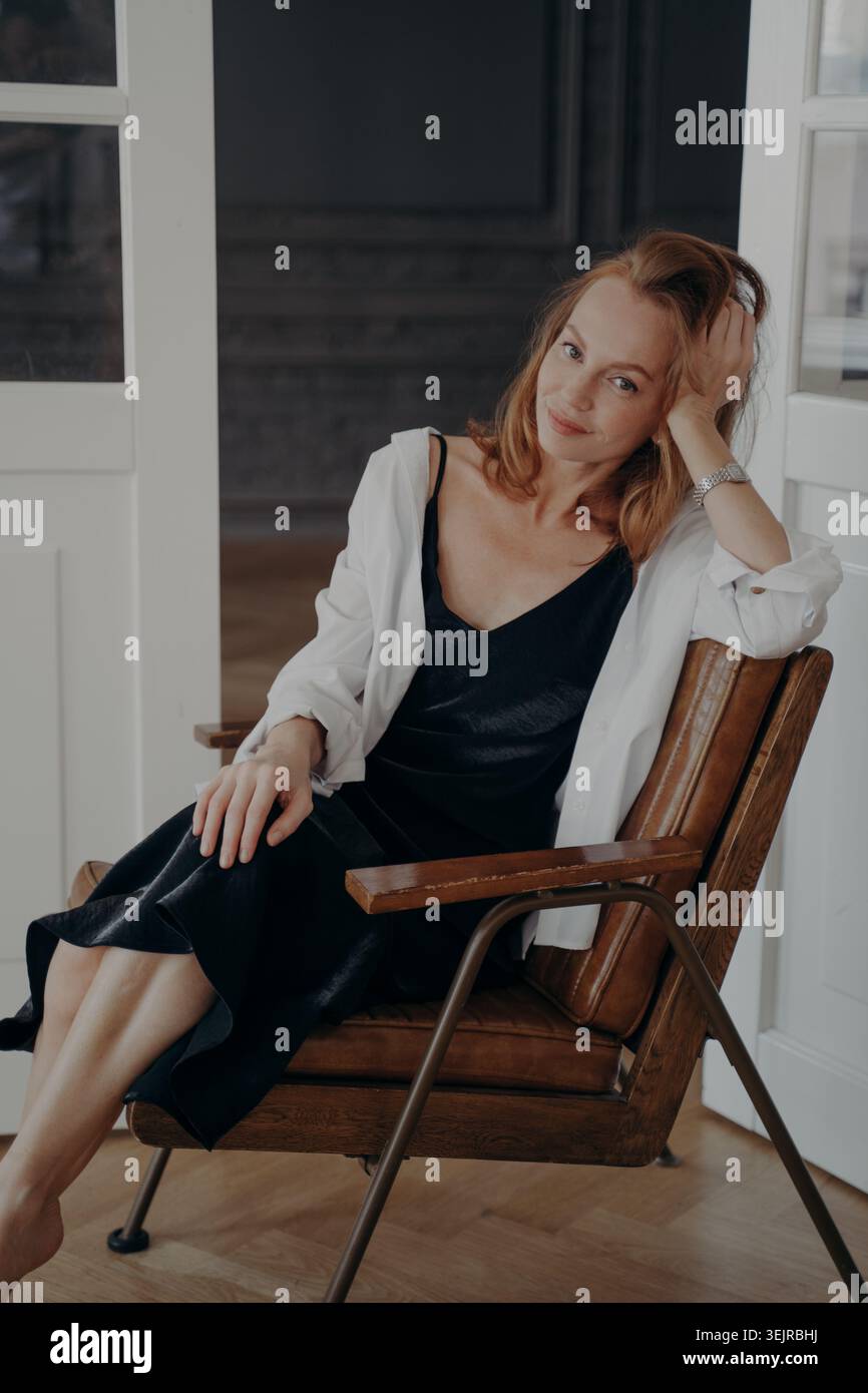 Donna dai capelli rossi seduta su una sedia di legno a casa con abito nero e camicia bianca Foto Stock