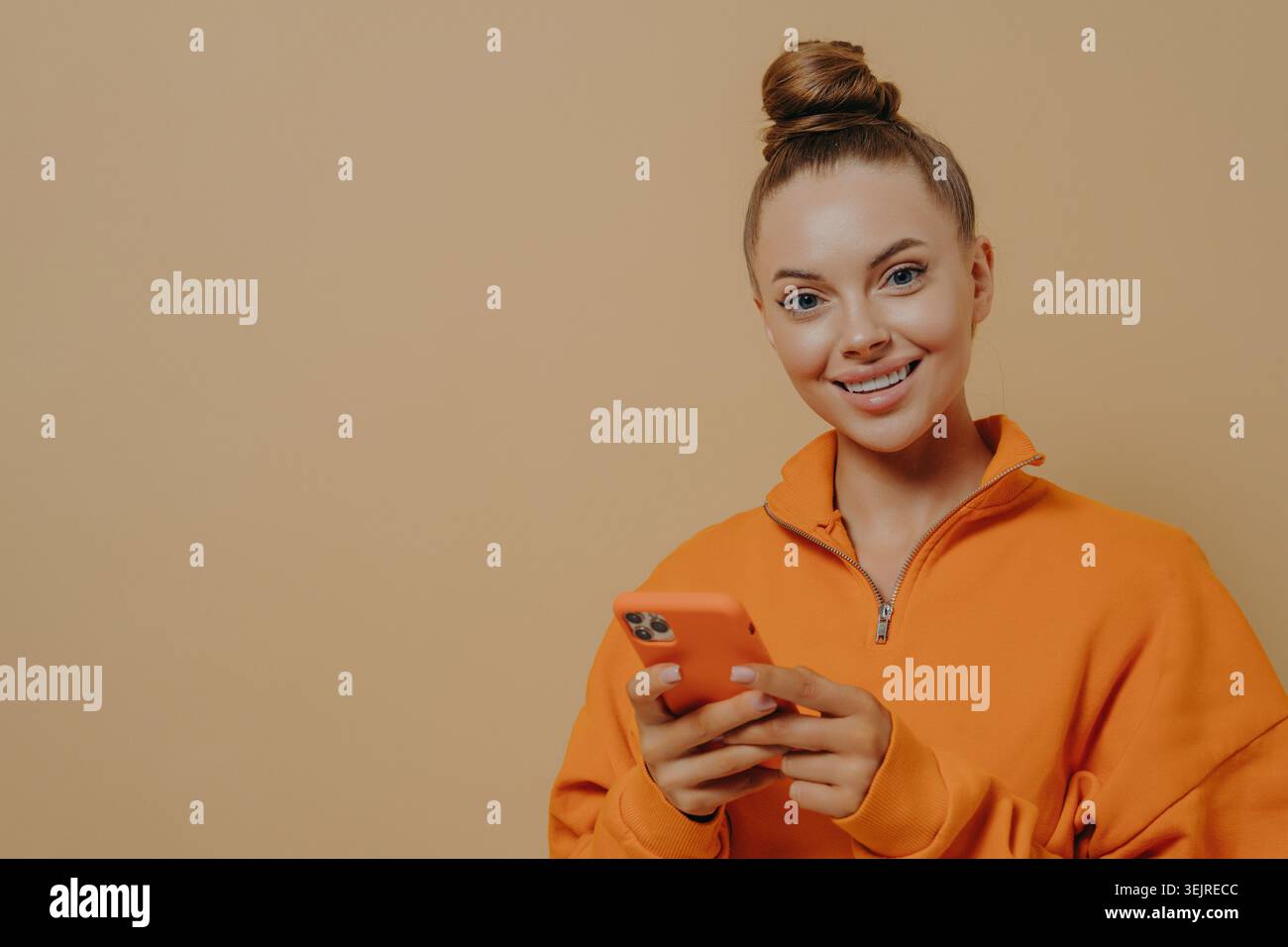 Allegra donna di generazione Z che utilizza lo smartphone sullo sfondo di Beige Studio indossando una felpa arancione e sorridendo durante la messaggistica Foto Stock
