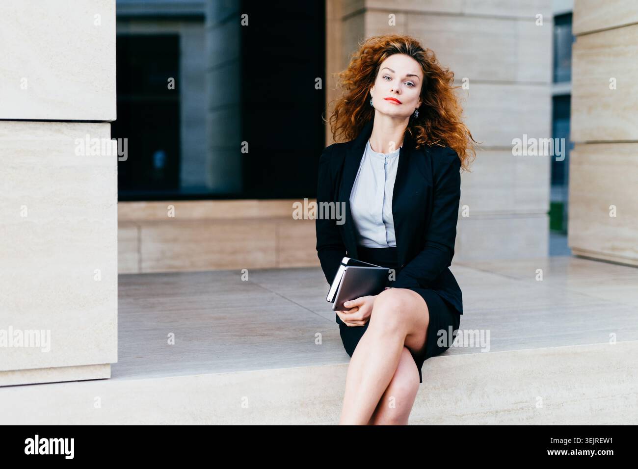Donna d'affari sicura in abito nero seduto con un tablet all'esterno di un moderno edificio per uffici Foto Stock