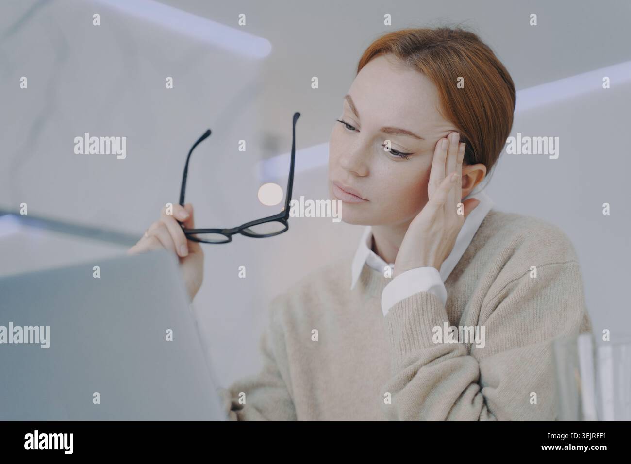 Una donna d'affari stanca tiene gli occhiali mentre lavora con un computer portatile in un ufficio moderno Foto Stock