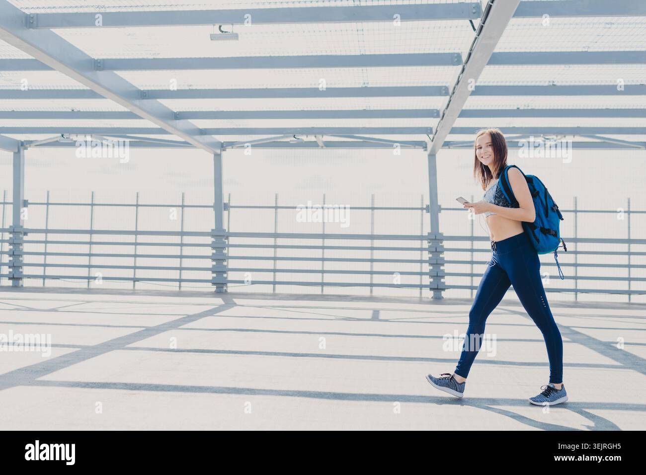 Una giovane donna con zaino attraversa un luminoso ponte urbano che regge lo smartphone Foto Stock