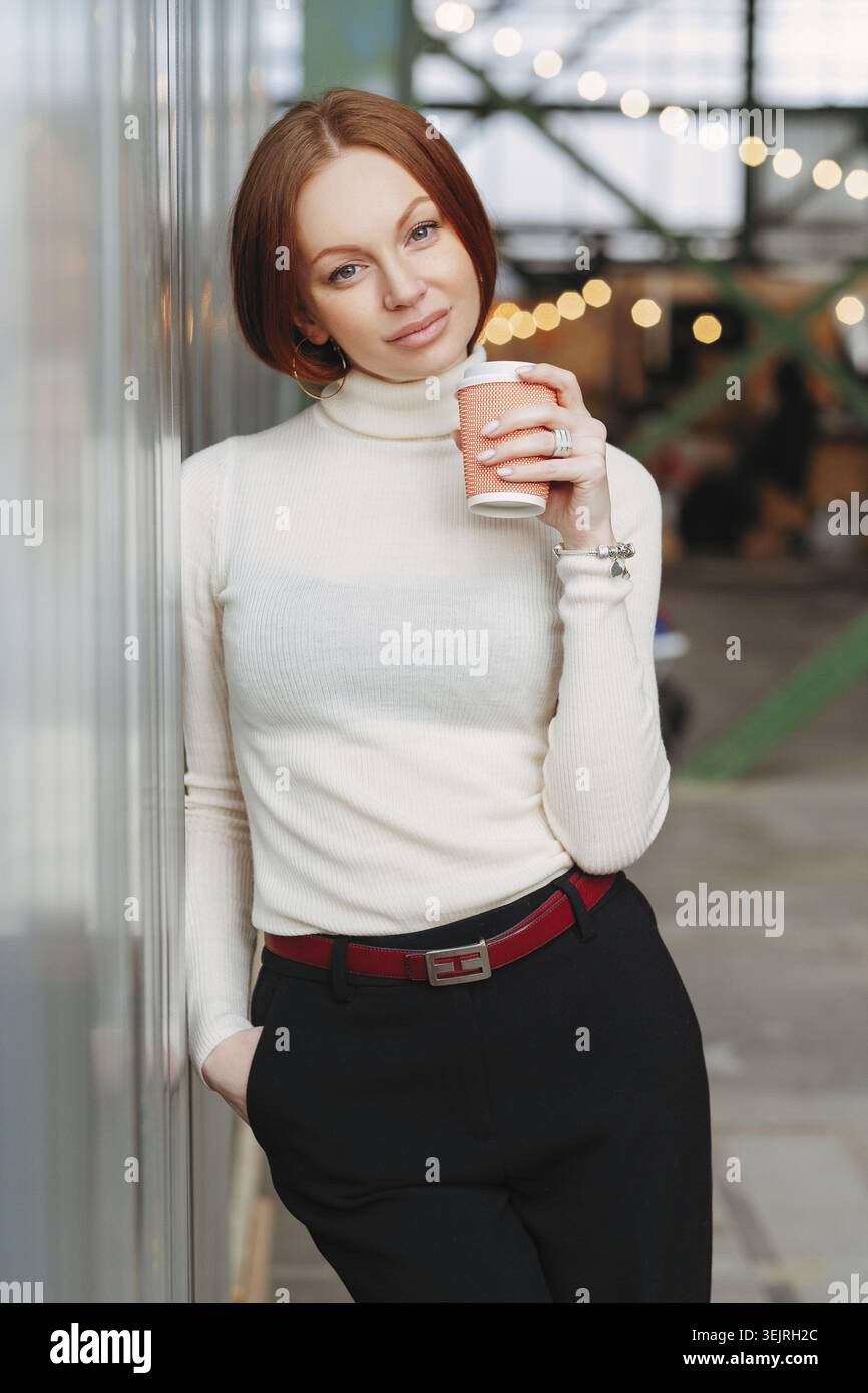 Donna rossa che tiene in mano una tazza di caffè da asporto mentre si appoggia alla finestra del caffè Foto Stock