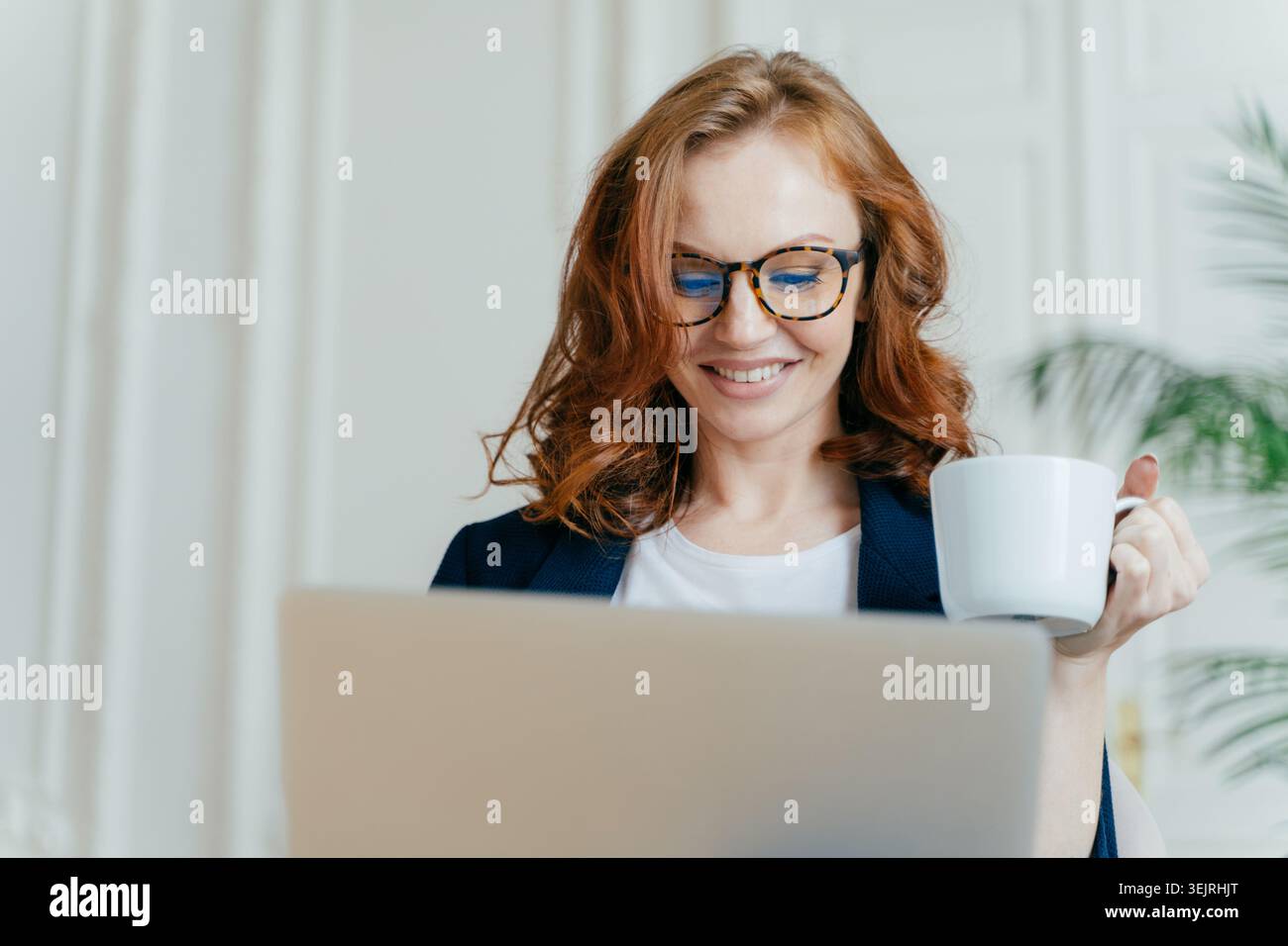Una donna d'affari dai capelli rossi sorride al computer portatile mentre tiene una tazza di caffè in ufficio Foto Stock