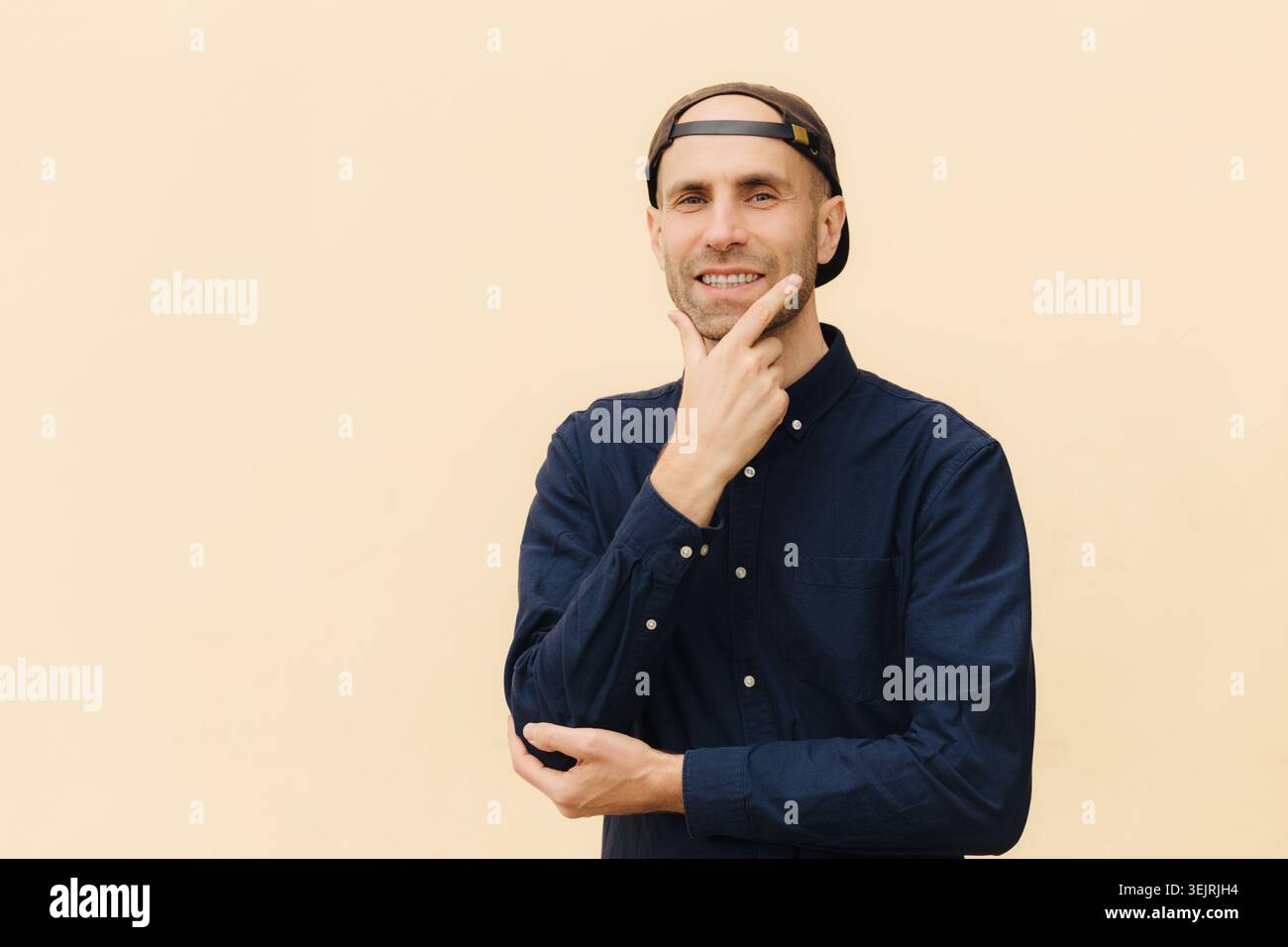 Un uomo casual sorridente con un cappellino da baseball all'indietro tocca il mento contro il muro beige Foto Stock