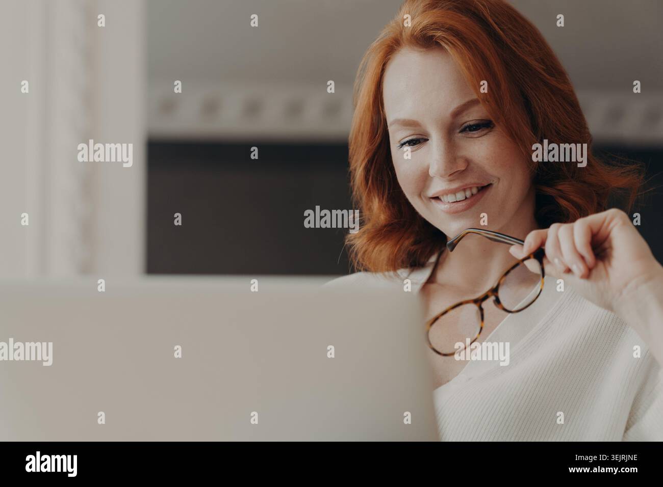 Una donna dai capelli rossi sorride mentre lavora con un laptop e tiene gli occhiali a casa Foto Stock