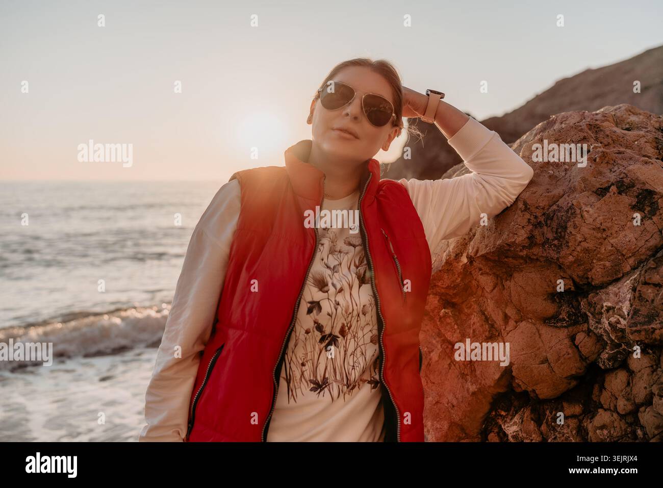 Donna tramonto spiaggia giovane donna che indossa giubbotto rosso e occhiali da sole appoggiati su una roccia vicino al mare durante l'ora d'oro Foto Stock