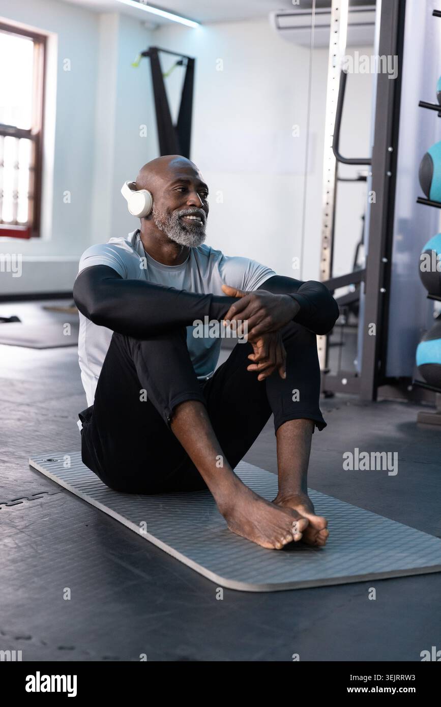 Senior afroamericano seduto su un tappetino grigio in palestra, indossando cuffie bianche, riposando Foto Stock