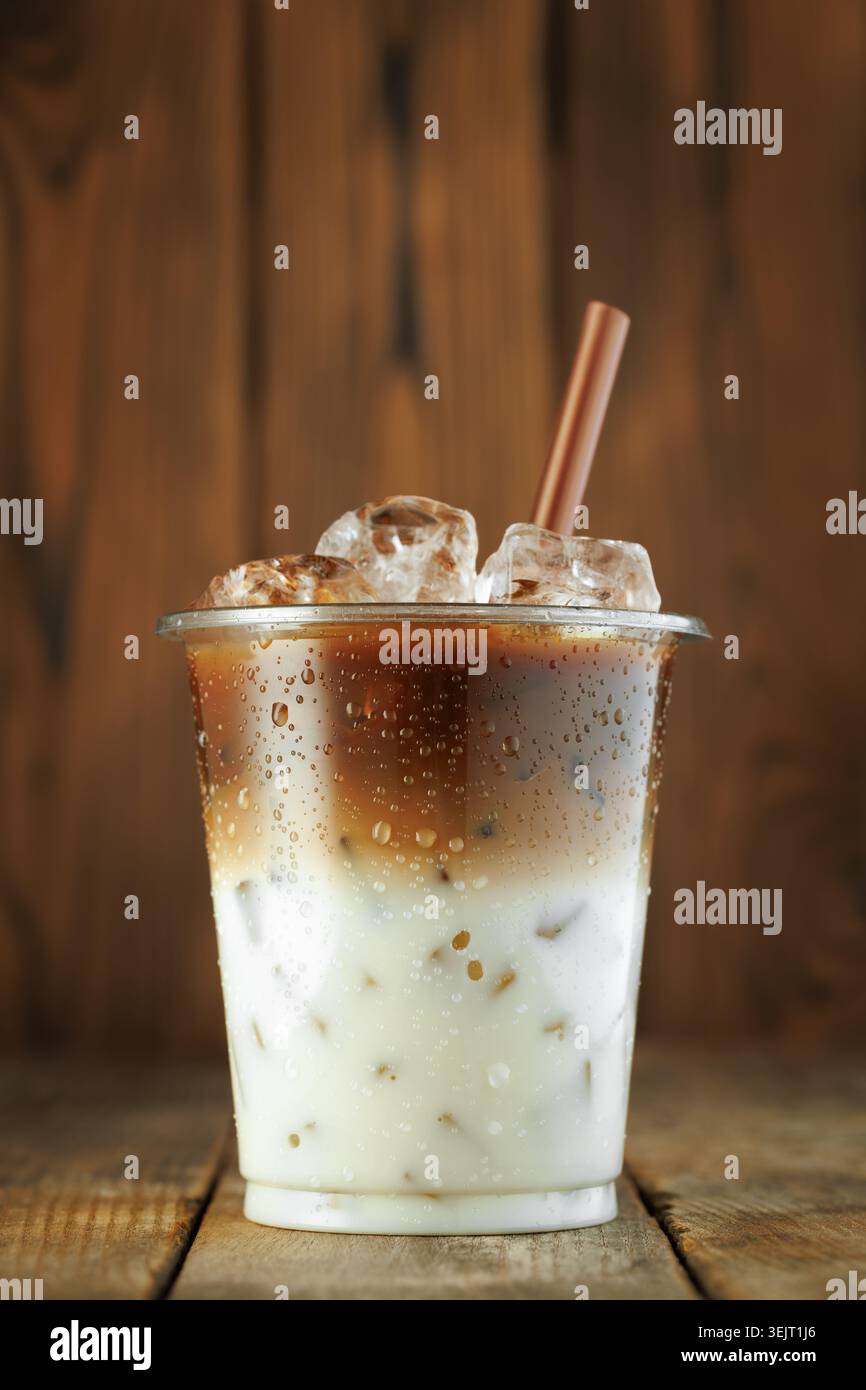 bevanda a base di latte ghiacciato in una tazza di plastica trasparente con paglia marrone, cubetti di ghiaccio e condensa, sempre su fondo in legno Foto Stock